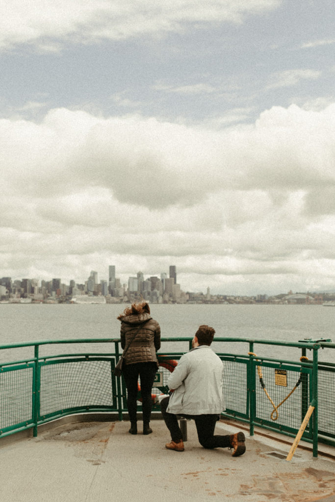 A surprise proposal on the ferry. The woman is facing away, looking at the water. The man is behind her, on one knee.