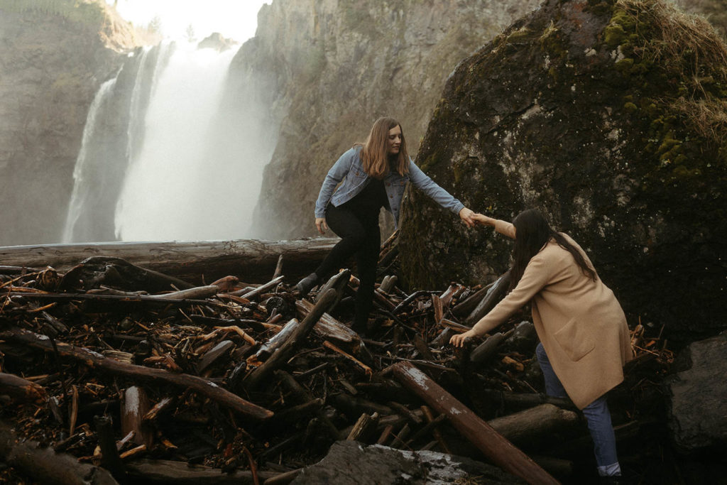 A couple is standing in front of Snoqualmie falls. One woman is standing on top of some logs, extending her hand to help the other woman climb up.