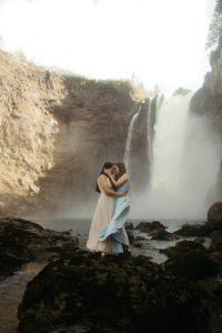 A couple is standing in front of Snoqualmie falls, facing each other. They have their foreheads together, with the waterfall behind them.