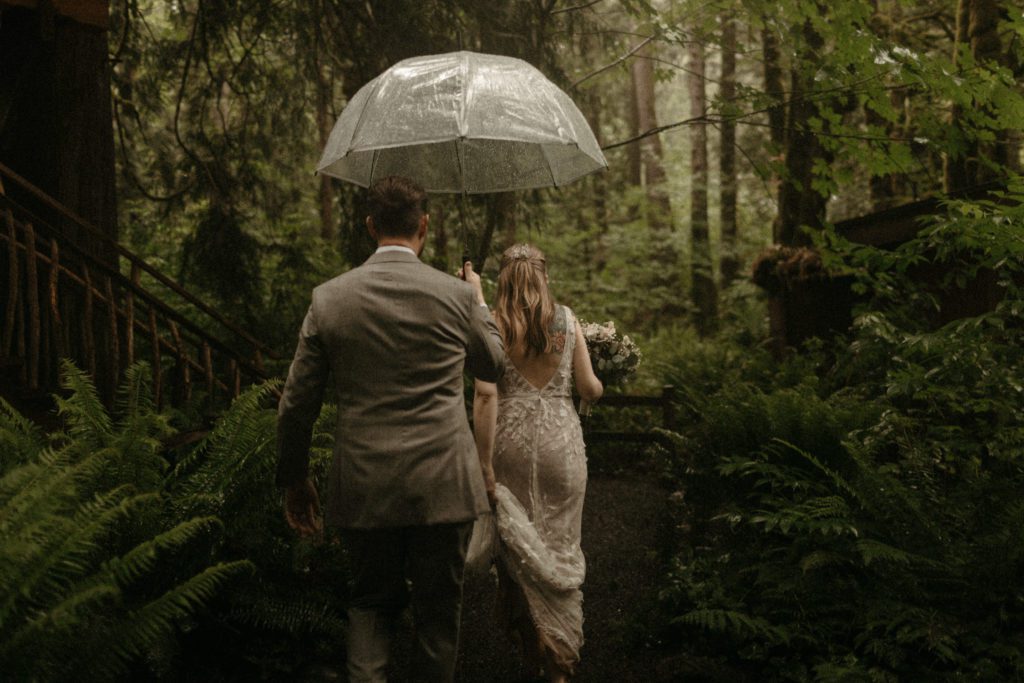 A couple walking through the forest, holding a clear umbrella at the Treehouse Point wedding venue.